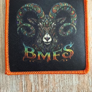 May include: A square patch with a black background and a bright orange stitched border. The center features a detailed illustration of a ram head with multicolored, intricate patterns on its horns. Below the ram, the letters BMFS are printed in a vibrant, rainbow-colored gradient font.