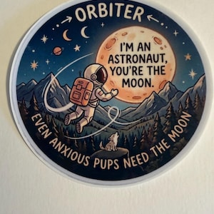 May include: A circular sticker featuring an astronaut floating in space toward a large moon. The text reads ORBITER, I'M AN ASTRONAUT, YOU'RE THE MOON, and EVEN ANXIOUS PUPS NEED THE MOON. A small dog sits on a mountain peak below the astronaut.