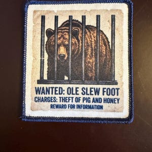 May include: A square embroidered patch with a dark blue border featuring a brown bear behind black vertical bars. The text reads: WANTED: OLE SLEW FOOT, CHARGES: THEFT OF PIG AND HONEY, REWARD FOR INFORMATION, printed on a light beige background.