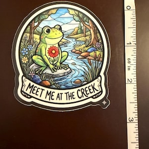 May include: A sticker featuring a green frog with a red flower on its chest, sitting on a rock in a creek. The sticker has a circular design with a forest and water scene. The text "MEET ME AT THE CREEK" is displayed on a banner. The sticker is approximately 3 inches in height.
