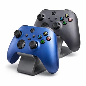 Könnte beinhalten: Zwei kabellose Gamecontroller, einer blau und einer grau, werden auf grauen Ständern präsentiert. Die Controller haben schwarze Tasten und farbige Aktionstasten. Das Xbox-Logo ist oben auf jedem Controller sichtbar.