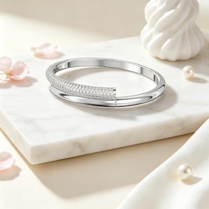 May include: A silver-toned hinged bangle bracelet featuring a polished finish on one half and a pavé-set section encrusted with numerous small, clear, sparkling stones on the other half. The bracelet has a sleek, circular design with a visible hinge.