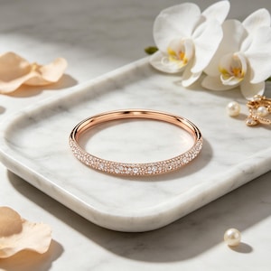 May include: A rose gold bangle bracelet featuring a pavé setting of small, clear, sparkling crystals along the outer surface. The bracelet has a smooth, polished rose gold finish on the inner band and a hinged clasp mechanism for closure.