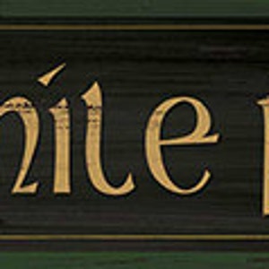 May include: A rectangular wooden sign with the Gaelic phrase "céad míle fáilte" in gold lettering. The sign has a dark green background with a distressed look, and a lighter green border.