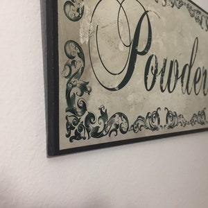 May include: A black framed sign with a white background and green decorative flourishes. The sign reads "Powder Room" in a decorative script font.
