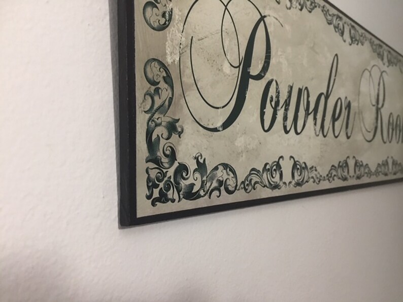 May include: A black framed sign with a white background and green decorative flourishes. The sign reads "Powder Room" in a decorative script font.