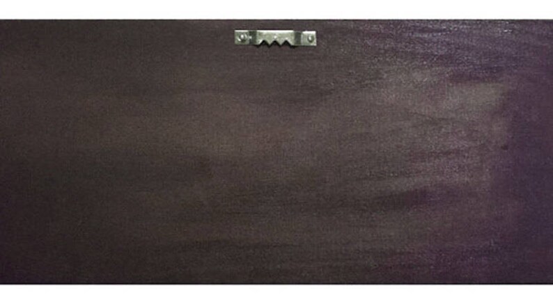 May include: Back of a rectangular, dark brown wall art piece with a metal sawtooth hanger attached at the top. The surface has a textured appearance with subtle variations in color.