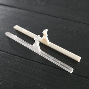 Könnte beinhalten: Zwei kleine, rechteckige Kunststoffobjekte, weiß und transparent, mit einem kleinen, erhabenen Dekorationselement. Die Objekte liegen auf einer dunklen Holzoberfläche.