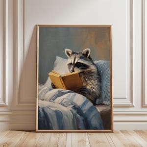 Puede incluir: Una impresión de arte mural enmarcada que representa a un mapache de pelaje blanco y negro sentado en la cama, apoyado contra almohadas y sosteniendo un libro abierto de color tostado. El mapache está cubierto por una manta a rayas azules.
