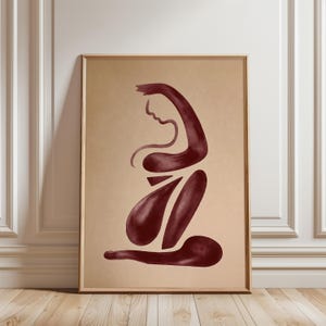 May include: A rectangular wall art print in a light wooden frame featuring a dark burgundy abstract figure on a solid beige background. The artwork displays a stylised, fluid silhouette of a seated person with curved lines and solid shapes.