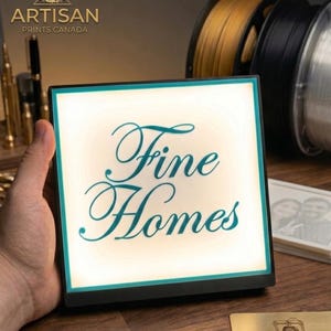 May include: A square, illuminated sign with the words "Fine Homes" in teal script. The sign is held in a hand, with a black frame and a white background. The Artisan Prints Canada logo is visible in the top left corner.