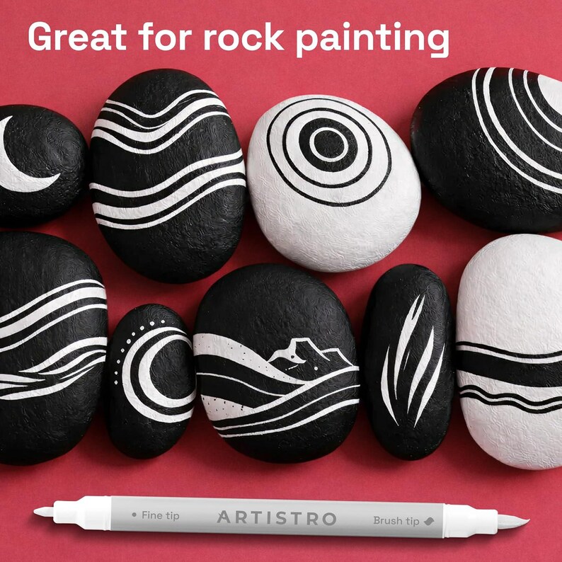 May include: A collection of smooth river stones painted with black and white geometric patterns, including waves, circles, and mountain silhouettes. A white dual-tip marker with the text ARTISTRO is positioned below the stones. The text Great for rock painting is visible.