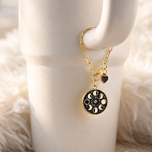 May include: A gold-colored necklace with a circular pendant featuring phases of the moon. The pendant is black with white and gold accents. A small black heart charm hangs near the clasp. The necklace is displayed on a white mug.