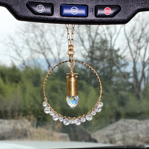 May include: A gold-colored car charm featuring a brass bullet casing with a faceted crystal at the base, suspended within a gold wire hoop. Clear, round crystals are attached to the bottom of the hoop. The charm hangs from a gold chain.