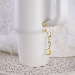May include: A gold-colored charm bracelet with celestial-themed charms. The bracelet features a small star, a rectangular blue gemstone, and a crescent moon charm. The bracelet is hanging on the handle of a white mug.