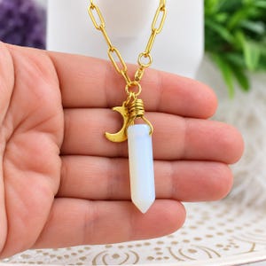 May include: A gold-toned chain necklace featuring a pendant with a milky white, cylindrical, pointed crystal stone wrapped in gold wire, accompanied by a small gold crescent moon charm hanging from the same circular link attachment.