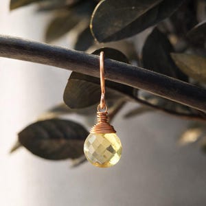 May include: A single earring with a faceted, yellow gemstone pendant. The pendant is wire-wrapped in copper and hangs from a copper ear wire. The earring is displayed against a blurred background of leaves and a dark branch.
