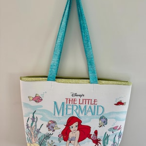 May include: White tote bag featuring Disney's The Little Mermaid. The bag has a colorful illustration of Ariel and sea creatures. It has turquoise handles and a green trim. The bag is designed for carrying items.