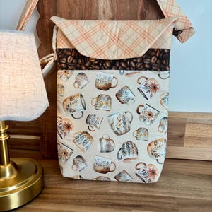May include: A quilted crossbody bag featuring a cream-colored fabric with a repeating pattern of various coffee mugs, a band of dark brown coffee beans, and a light orange plaid flap and strap. The bag has a rectangular shape and a fold-over closure.