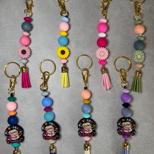 May include: A collection of beaded keychains with gold-colored hardware. Each keychain features a variety of colorful beads, decorative flower accents, and a tassel. Some keychains have a charm that reads "My Weekend is Booked."