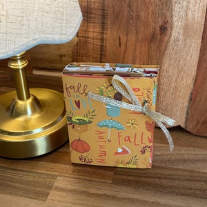 May include: A small, rectangular, mustard-yellow paper booklet featuring autumn-themed illustrations of pumpkins, sunflowers, and umbrellas. The text reads "fall IN LOVE" and "Autumn". A silver glittery ribbon is tied around the center of the booklet.