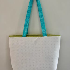 May include: A white tote bag with a dotted pattern and a green trim. The bag has turquoise blue handles. The bag is hanging against a white wall.