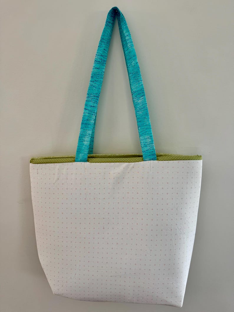 May include: A white tote bag with a dotted pattern and a green trim. The bag has turquoise blue handles. The bag is hanging against a white wall.