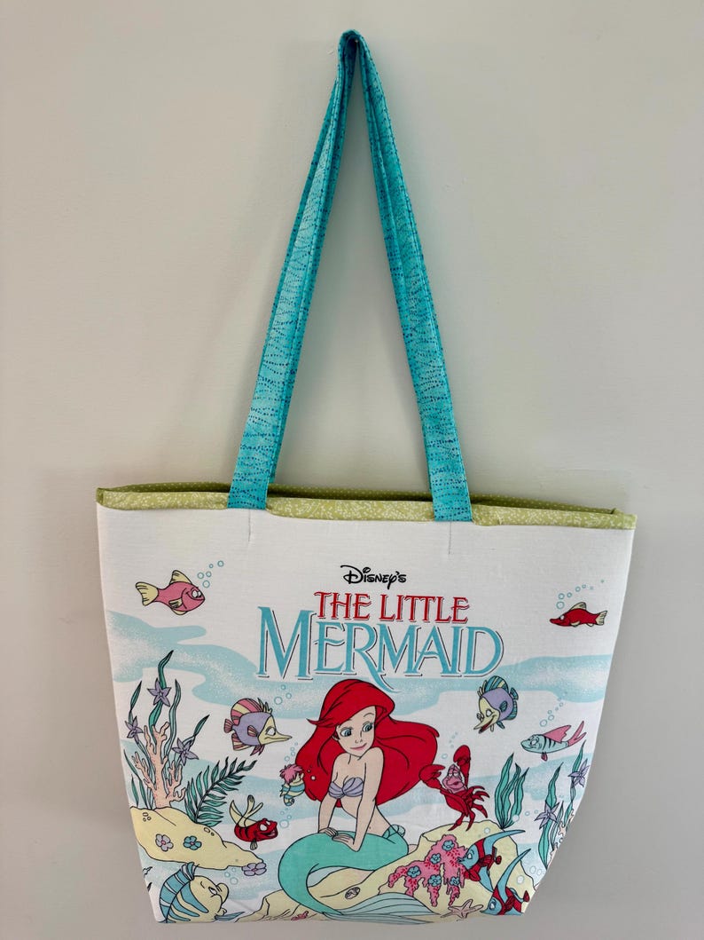 May include: White tote bag featuring Disney's The Little Mermaid. The bag has a colorful illustration of Ariel and sea creatures. It has turquoise handles and a green trim. The bag is designed for carrying items.