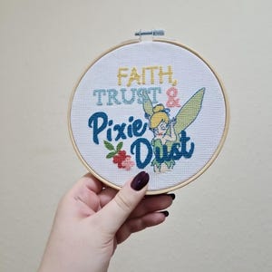 May include: A circular wooden embroidery hoop containing white fabric with a cross-stitch design. The text reads Faith, Trust & Pixie Dust in yellow, blue, and red. A small green and yellow fairy figure is stitched next to the word Dust.