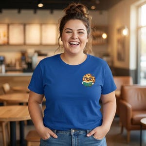 May include: A royal blue t-shirt featuring a cartoon cookie character wearing glasses and reading a book. The cookie has a chocolate chip design and is holding a green book. The person is wearing blue jeans.