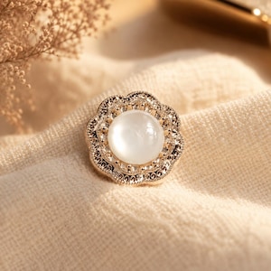 May include: A gold-toned, flower-shaped button with a central, round, white, opalescent stone. The button's edge is detailed with small, clear crystals and intricate filigree. The button is set against a cream-colored fabric.