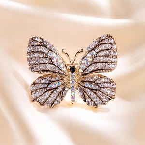 May include: A gold-toned butterfly brooch featuring wings encrusted with clear, iridescent rhinestones. The central body of the butterfly is adorned with a single black stone at the top, followed by a vertical row of smaller, shimmering iridescent stones.