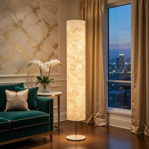May include: A tall, cylindrical floor lamp featuring a textured, off-white fabric shade that emits a warm, diffused glow. The lamp is supported by a slender, polished silver-tone metal pole and a circular, white marble-patterned base.