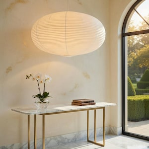 May include: A large, oval-shaped white paper pendant light fixture with horizontal ribbed detailing hangs from a thin cord above a rectangular console table featuring a white marble top and a gold-toned metal frame with vertical support legs.