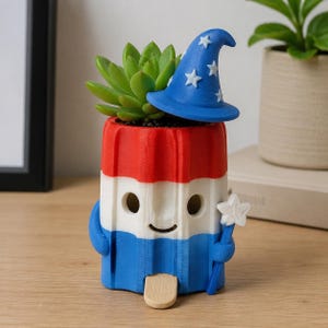 May include: A cylindrical planter shaped like a red, white, and blue ice lolly with a smiling face. It features a blue wizard hat with white stars, a small white star-shaped wand, and a wooden stick at the base. A green succulent plant sits inside.