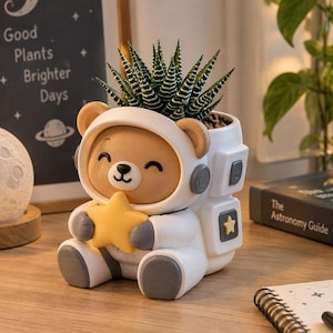 May include: A white and brown resin planter shaped like a bear wearing an astronaut suit, holding a yellow star. The bear has a smiling face and grey accents on the suit. A green succulent plant is potted inside the top of the astronaut helmet.