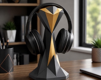 May include: A black and gold geometric headphone stand holding a pair of black over-ear headphones. The stand features a faceted, angular design with a wide base and a central pillar that tapers upward, showcasing contrasting matte black and gold surfaces.