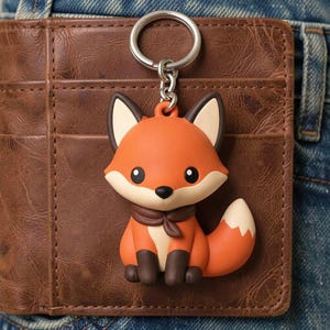 May include: A cute orange fox keychain with a white belly, ears, and tail tip. The fox has a brown scarf and dark brown feet. The keychain is attached to a silver metal ring and chain. The fox is resting on a brown leather wallet.