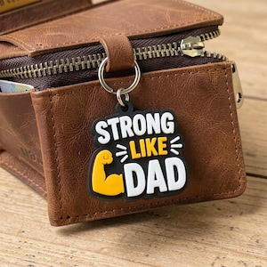 May include: A brown leather wallet with a zipper closure and a keychain. The keychain is black with white and yellow text that reads "STRONG LIKE DAD" and features a cartoon arm flexing.