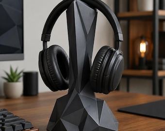 May include: A matte black geometric headphone stand with a faceted, angular design. The stand features a wide, stable base that tapers upward to support a pair of over-ear black headphones with cushioned ear cups resting on the top section.