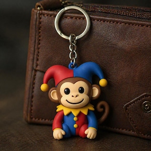 May include: A colorful monkey keychain with a jester hat in red and blue. The monkey has a brown face, ears, and tail, with a yellow collar and a smiling expression. The keychain is attached to a silver metal ring.