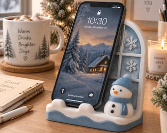 May include: A smartphone resting on a white and blue resin phone stand shaped like a window frame with a small snowman wearing a blue hat and scarf. The stand features two white snowflake accents and a wavy base, holding the phone upright.