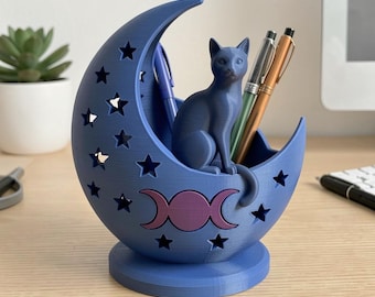 May include: A blue crescent moon-shaped desk organizer with a cat figurine on top. The organizer has star cutouts and a triple moon symbol. Pens are placed inside the organizer. The desk organizer is a unique and decorative item.