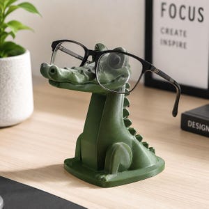 May include: A dark green crocodile-shaped eyeglass holder made of a smooth material. The crocodile is in a seated position with its snout extended forward, providing a resting place for a pair of black-framed eyeglasses that are perched on its nose.