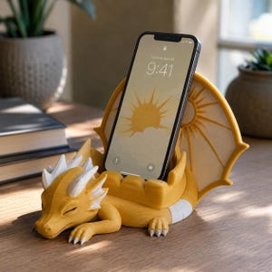 May include: A mustard yellow dragon-shaped smartphone stand featuring white horns and a large wing backdrop. The dragon is in a resting position with its eyes closed, providing a stable base for a smartphone displaying a sun graphic and the time 9:41.