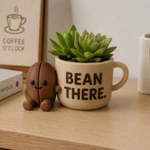 Puede incluir: Una maceta de cerámica beige con forma de taza y el texto BEAN THERE. impreso en marrón oscuro, que contiene una suculenta verde. Junto a la taza hay una pequeña figura marrón con forma de grano de café y una cara sonriente.