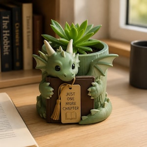 Puede incluir: Una maceta con forma de dragón verde claro que contiene una suculenta. El dragón tiene cuernos y alas de color crema y sostiene un libro marrón con una etiqueta beige que dice "JUST ONE MORE CHAPTER" junto a un icono de un libro abierto.