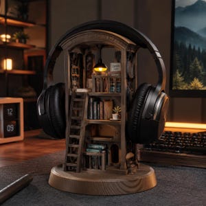 Puede incluir: Un soporte de madera para auriculares con forma de biblioteca de varios niveles, con una escalera en miniatura, estantes llenos de libros, una pequeña planta en maceta y una lámpara colgante. Auriculares negros sobre el arco.
