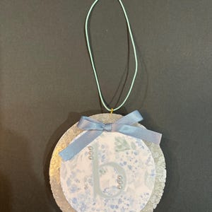 May include: A round, layered ornament with a floral design and the letter "b". The ornament features a light blue ribbon bow and a light blue string for hanging. Small silver beads accent the letter.