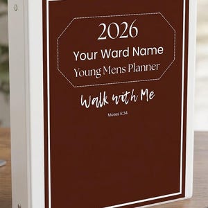 May include: A white binder with a brown cover featuring the text "2026 Your Ward Name Young Mens Planner Walk with Me Moses 6:34". A pen rests on the surface next to the binder.